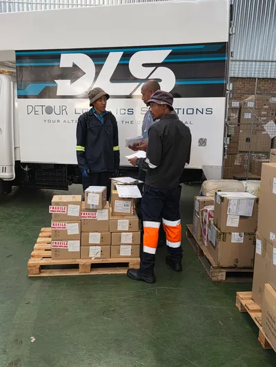 Two DLS staff checking paperwork beside pallets of boxes in front of a DLS truck in a warehouse.