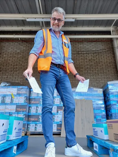 Sybrand Engelbrecht, owner of DLS, standing on pallets holding delivery paperwork, with stacked stock behind him.