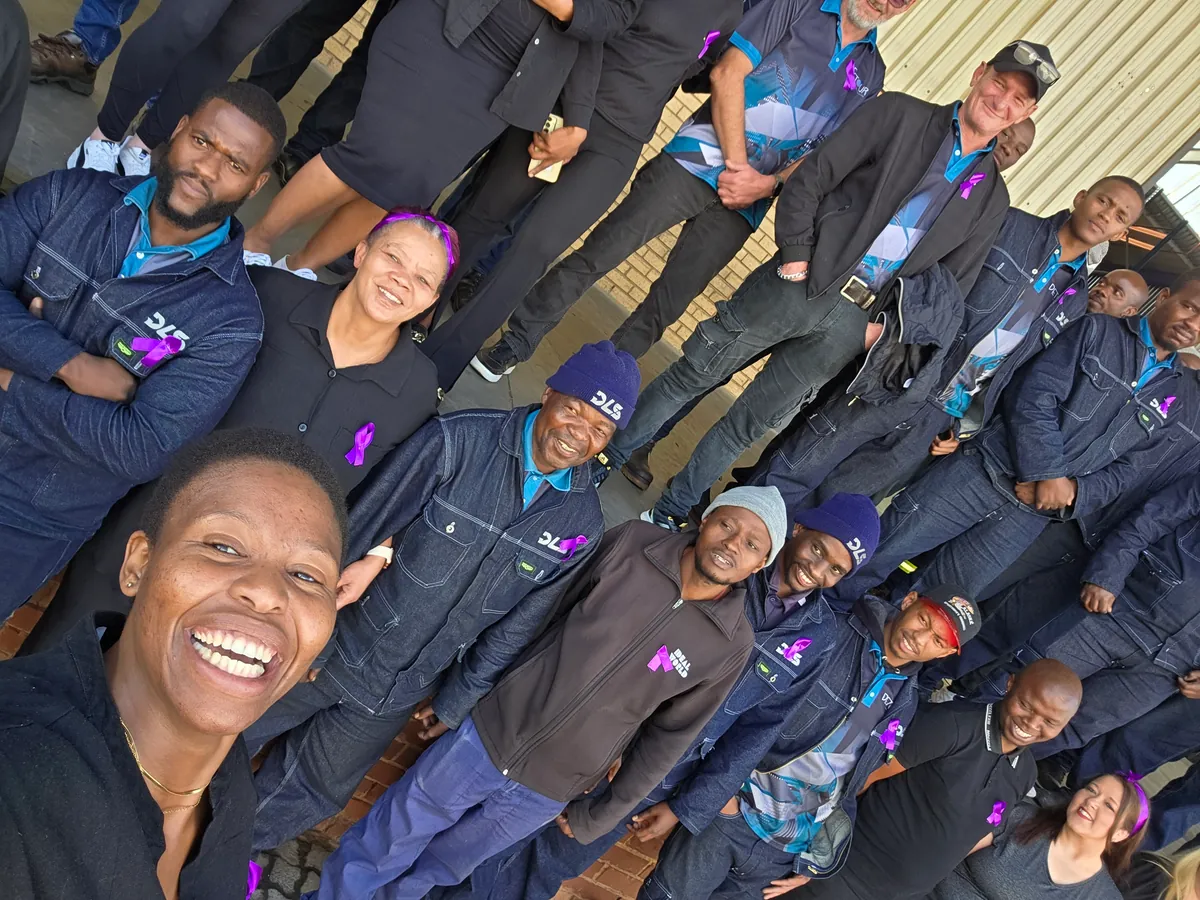 A team member's selfie with about fifteen DLS colleagues in denim workwear behind her, some wearing purple ribbons.