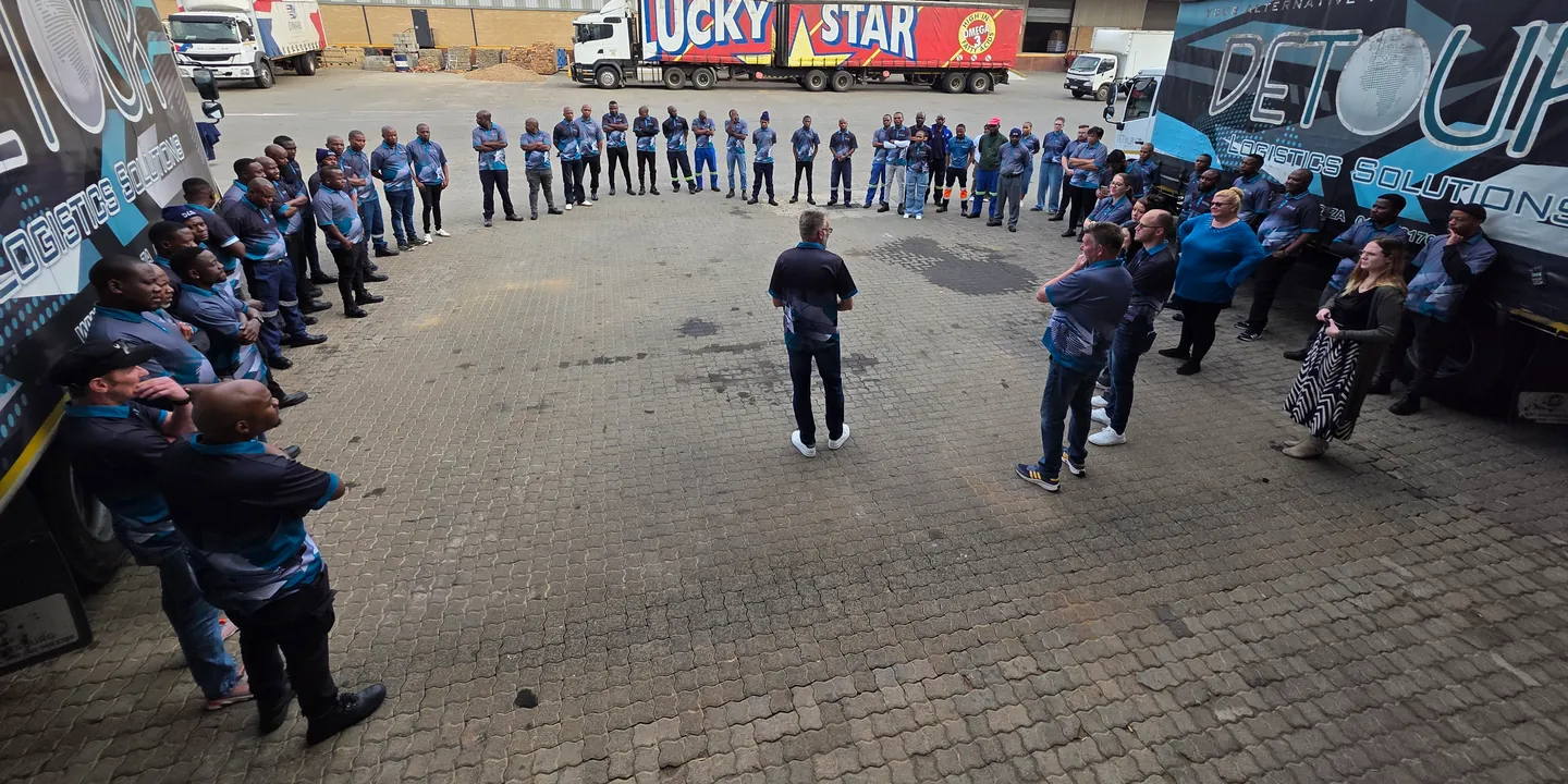 DLS staff in branded shirts stand in a wide circle on the paved yard between two DETOUR-liveried trucks, with one person in the centre facing them.